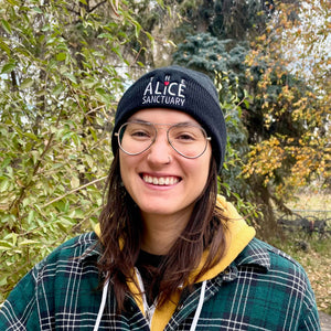 Smiling woman wearing black knitted cuff toque with the words,"The Alice Sanctuary," embroidered on the front.